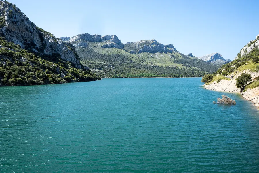 Blick vom steinigen Ufer über die ruhige, türkisfarbene Wasserfläche des Gorg Blau auf die Berge am gegenüberliegenden Ende.