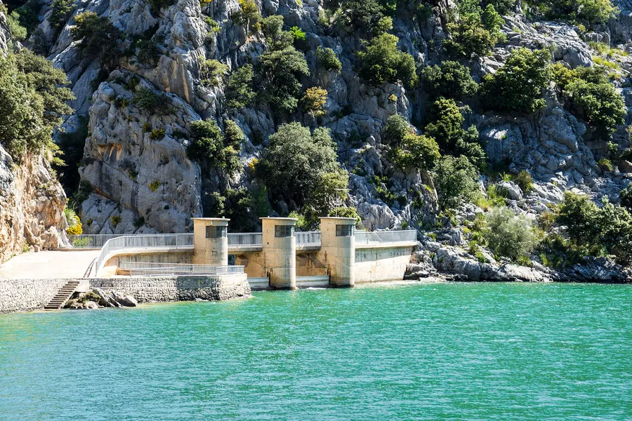 Die Staumauer des Gorg Blau mit Schleusentoren aus Stahl, davor das türkisfarbene Wasser, dahinter die Felswand.