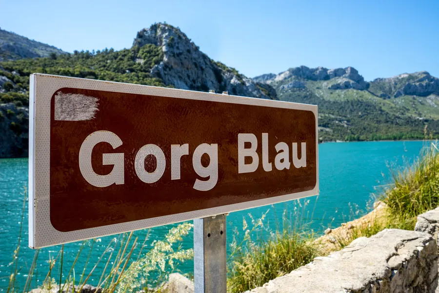 Braunes Hinweisschild mit der Aufschrift Gorg Blau am Straßenrand der MA-10, dahinter der See und die Berge.