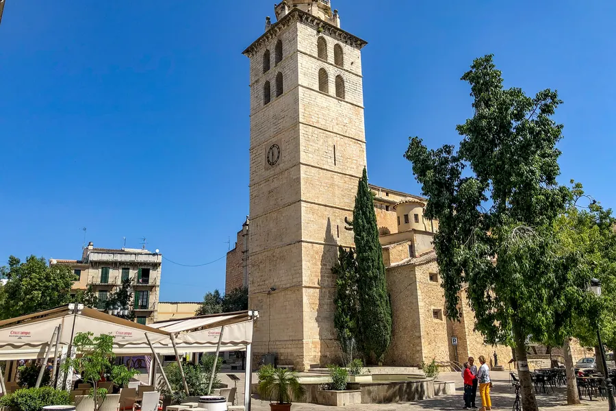 Der hohe helle Glockenturm der Pfarrkirche Santa Maria la Major in Inca mit Uhr und offenem Glockengeschoss, davor Bäume und Zypressen.
