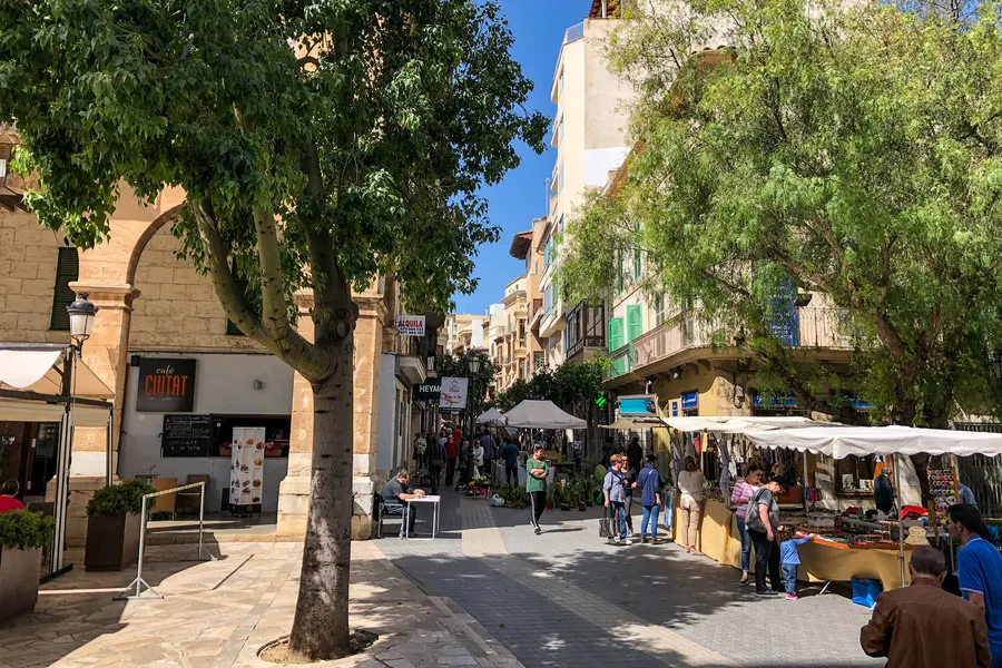 Marktstände mit weißen Planen in einer Fußgängerstraße von Inca, dazwischen Passanten unter großen Bäumen.