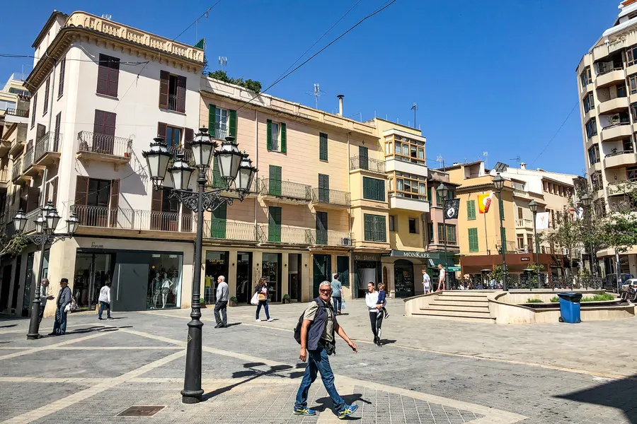 Belebte Fußgängerzone in Inca mit hohen Straßenlaternen, Wohn- und Geschäftshäusern und Passanten am Vormittag.
