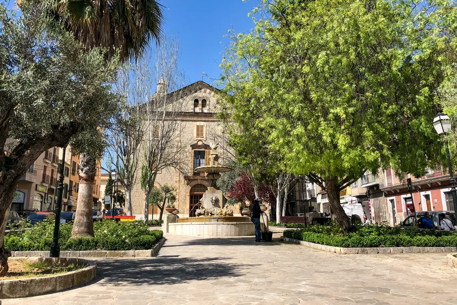 Schattiger Platz in Inca mit großen alten Bäumen, gepflastertem Boden und einem Steingebäude im Hintergrund.
