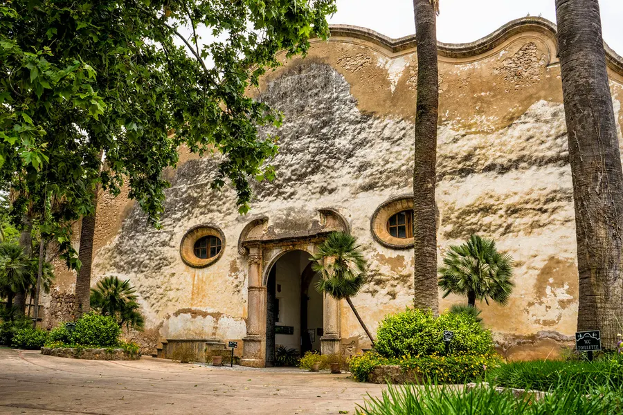Geschwungene alte Mauer des Anwesens Alfàbia mit Rundfenstern und Rundbogentor, davor Palmen und ein gekiester Vorplatz.

