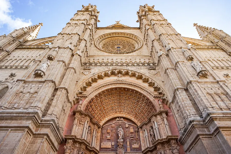Das Hauptportal der Kathedrale von Palma von unten fotografiert, mit hohem Bogenfeld, reich verziertem Steinschmuck, Rosette und zwei Türmen.
