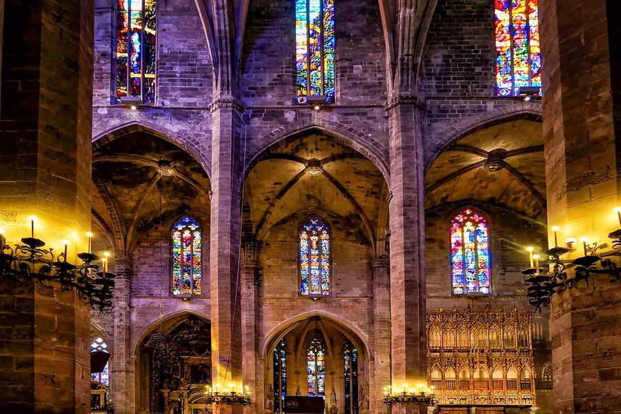 Blick in das Innere der Kathedrale von Palma mit hohen Spitzbogenarkaden, farbigen Glasfenstern und einem vergoldeten Retabel im Seitenschiff.