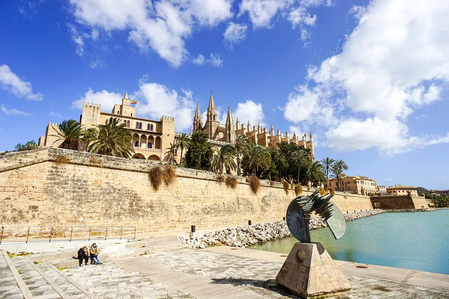 Die Kathedrale von Palma und der Palast Almudaina hinter der alten Stadtmauer mit Palmen, davor eine moderne Metallskulptur am Beckenrand.
