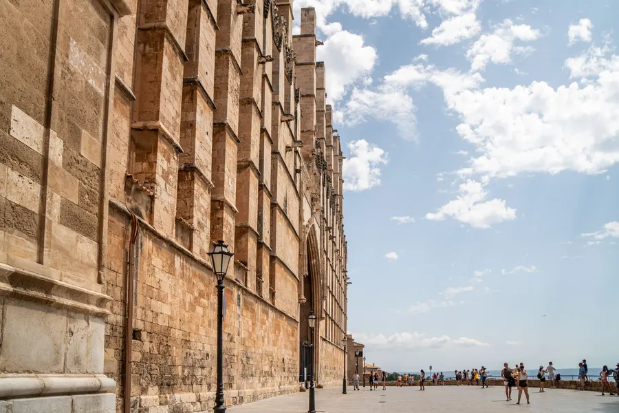 Die lange Südflanke der Kathedrale von Palma mit Strebepfeilern, davor die Promenade und das Meer.
