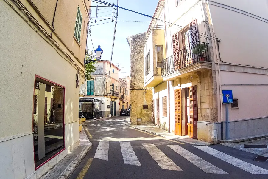 Ruhige Gasse in Llubí mit hellen Häusern, grünen Fensterläden und einem Zebrastreifen, im Hintergrund der Kirchturm.
