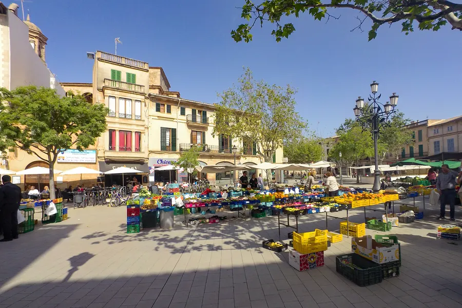 Marktstände mit Obst- und Gemüsekisten auf einem Platz in Llucmajor, dahinter Häuserzeilen und Bäume.