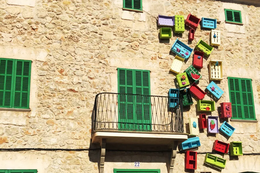 Bunt bemalte Holzkisten, die als Kunstwerk an der Natursteinfassade eines Hauses mit grünen Fensterläden befestigt sind.
