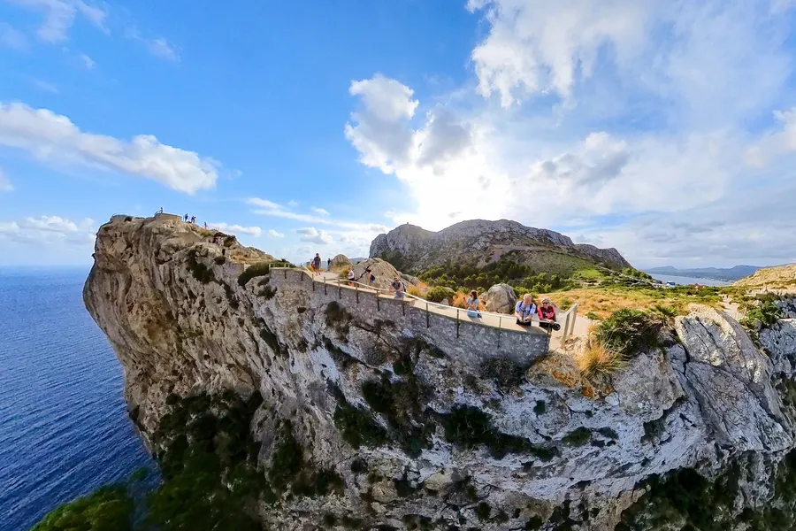 Gemauerter Weg mit Geländer, der am Rand einer senkrecht ins Meer abfallenden Felswand entlangführt; einzelne Besucher lehnen am Geländer, im Hintergrund der Bergkamm und in der Ferne die Bucht von Pollença.