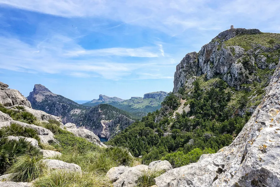 Blick über bewaldete Bergrücken der Halbinsel Formentor bis zu den fernen Felsspitzen am Kap; rechts oben auf einem Gipfel die kleine Silhouette des Wachturms von Albercutx, im Vordergrund helle Kalkfelsen und Zwergpalmen.
