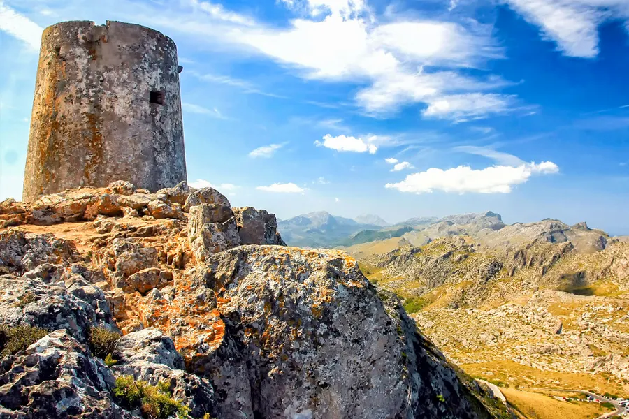 Runder, verwitterter Wachturm aus Naturstein auf einer Felskuppe, im Vordergrund flechtenbewachsene Kalkblöcke, dahinter die weit gestaffelten Bergketten der Serra de Tramuntana unter einem Himmel mit Schönwetterwolken.

