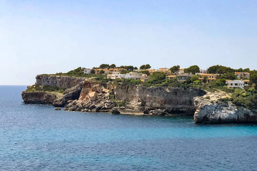 Steilküste bei Santanyí mit senkrechten hellen Felswänden über tiefblauem Wasser, oben auf der Kante einzelne weiße Häuser zwischen Kiefern.