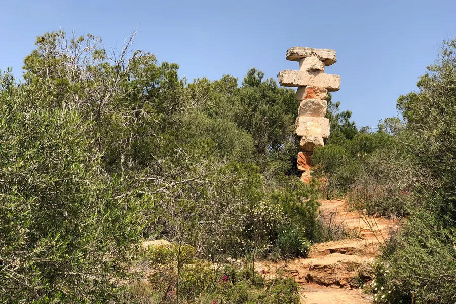 Schmaler Naturweg durch niedrige Macchia und Kiefern zum Aussichtspunkt, im Hintergrund die Steinskulptur und der Himmel.
