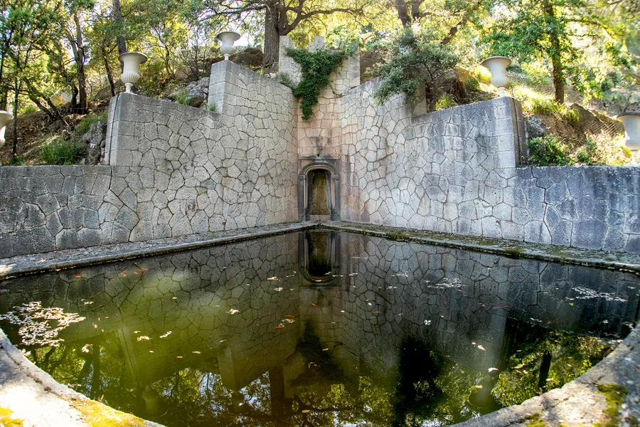 Rechteckiges steinernes Wasserbecken im Garten von Miramar, dahinter eine Mauer mit Nische und alte Bäume.