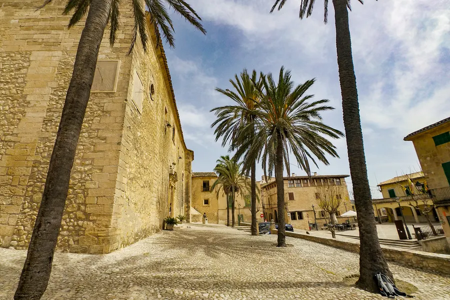 Gepflasterter Platz in Montuïri mit hohen Palmen, einer langen Steinfassade und den Häusern des Ortes im Hintergrund.
