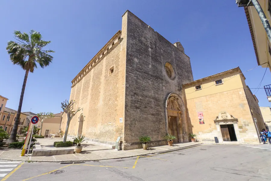 Die wuchtige spätgotische Fassade der Kirche Sant Joan Baptista in Muro mit hohem Portal, davor eine Palme und der leere Platz.