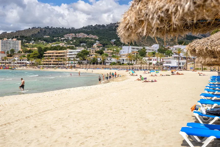 Weiter Sandstrand von Peguera mit Badegästen und Liegen, dahinter die Hotelzeile und die bewaldeten Hügel.
