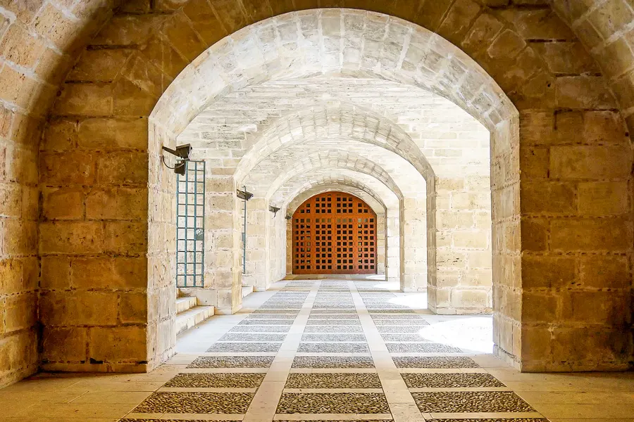 Langer tonnengewölbter Gang aus hellem Sandstein im Palau de l'Almudaina mit gemustertem Steinboden und einer Holztür am Ende.