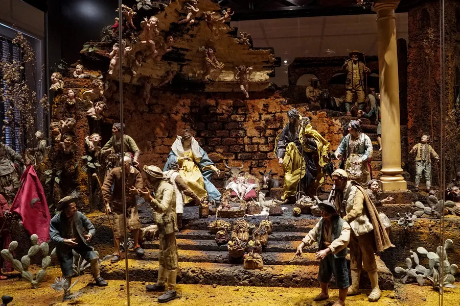 Gesamtansicht der neapolitanischen Krippe im Palau March: eine Felsenlandschaft mit gemauerter Ruine, darüber schwebende Engel, davor Hirten, Marktkörbe und die Heilige Familie.
