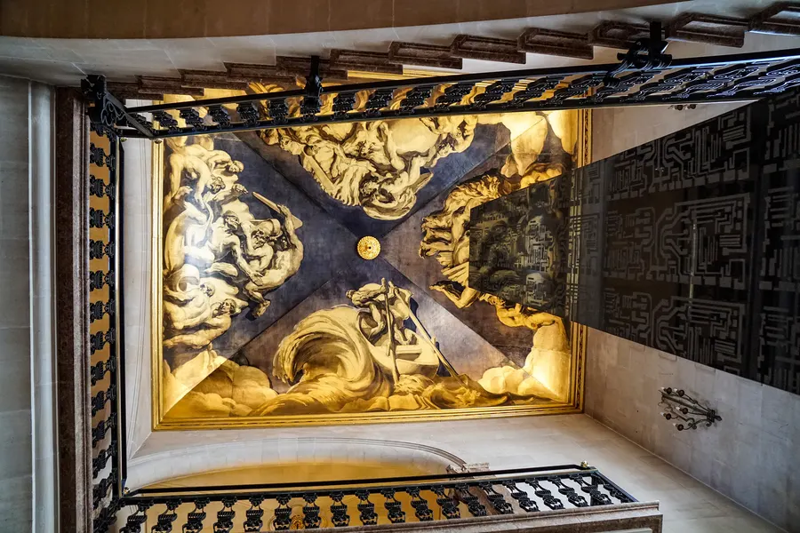 Blick nach oben in das Treppenhaus des Palau March: ein großes Deckengemälde in Gold- und Grautönen mit ringenden Figuren und Wolkenbändern, gerahmt von einem schmiedeeisernen Geländer.
