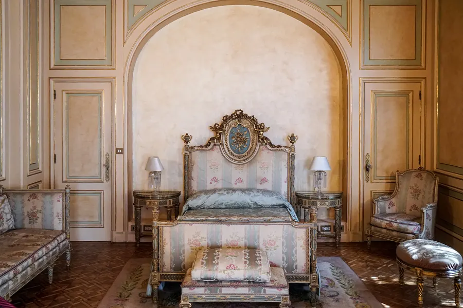 Ein Schlafzimmer im Palau March mit vergoldetem Bett in einer Wandnische, gestreiften Seidenbezügen, zwei Nachttischen und einem Sofa unter hellen Stuckpaneelen.