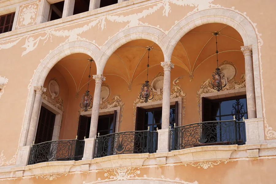 Die dreibogige Loggia an der Fassade des Palau March mit Marmorsäulen, schmiedeeisernen Balkongittern, Laternen und weißem Rankenwerk auf ockerfarbenem Putz.
