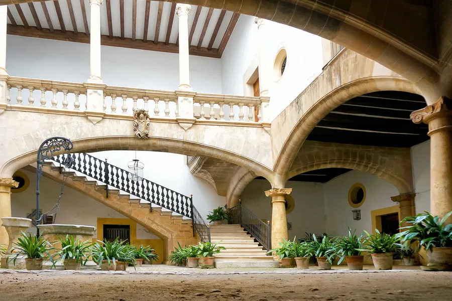 Herrschaftlicher Innenhof in Palma mit Rundbogenarkaden auf Säulen, einer geschwungenen Steintreppe und einem Balkon darüber.

