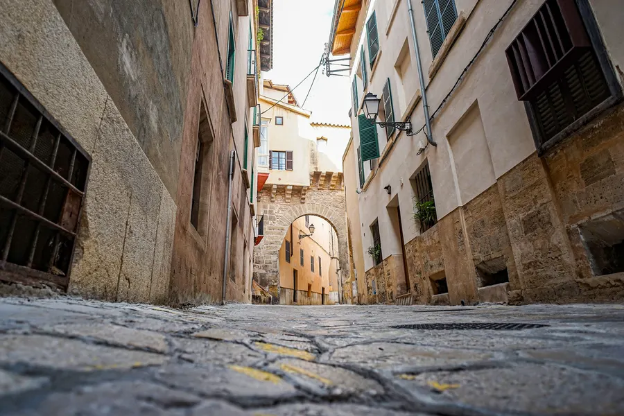 Enge gepflasterte Altstadtgasse in Palma zwischen hohen Häuserwänden, am Ende ein Torbogen mit Durchblick.
