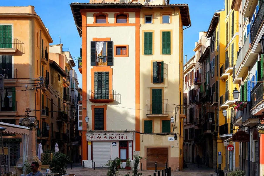 Schmales hohes Eckhaus an der Plaça des Coll in Palma mit grünen Fensterläden und bunten Nachbarfassaden.
