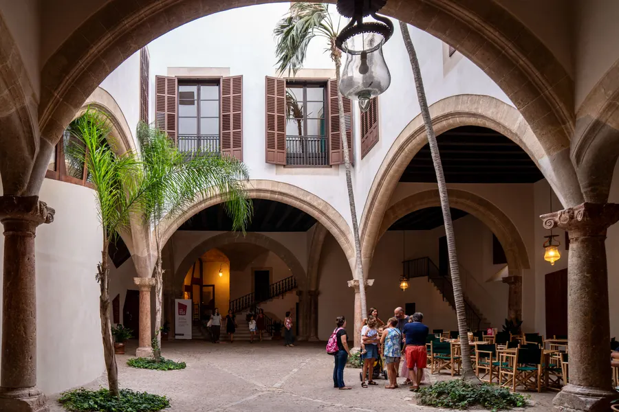 Lichter Innenhof des Can Balaguer in Palma mit Rundbogenarkaden auf Säulen, Palmen in Kübeln und Tischen eines Cafés.
