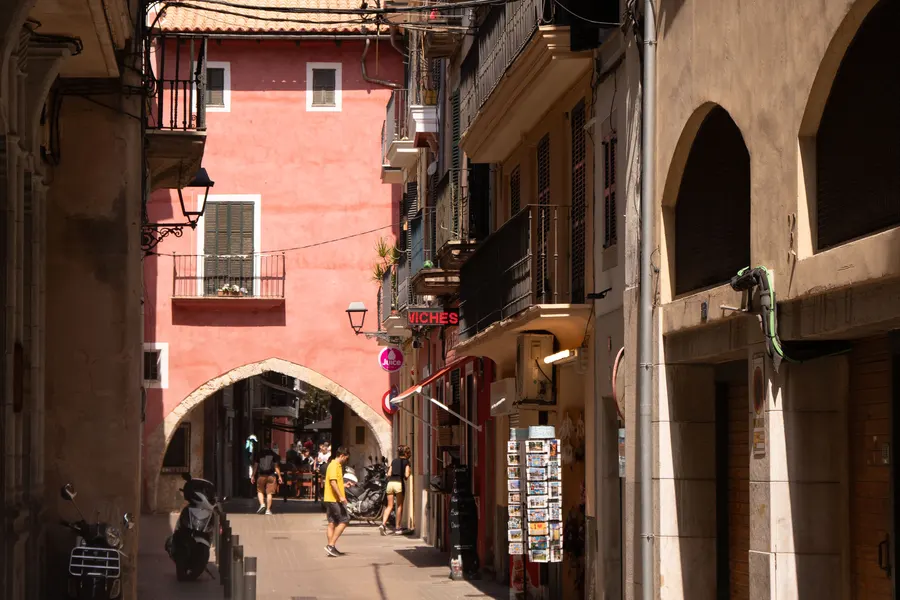 Gasse in Palmas Altstadt mit rot verputztem Haus, einem Torbogen und Ladenschildern.
