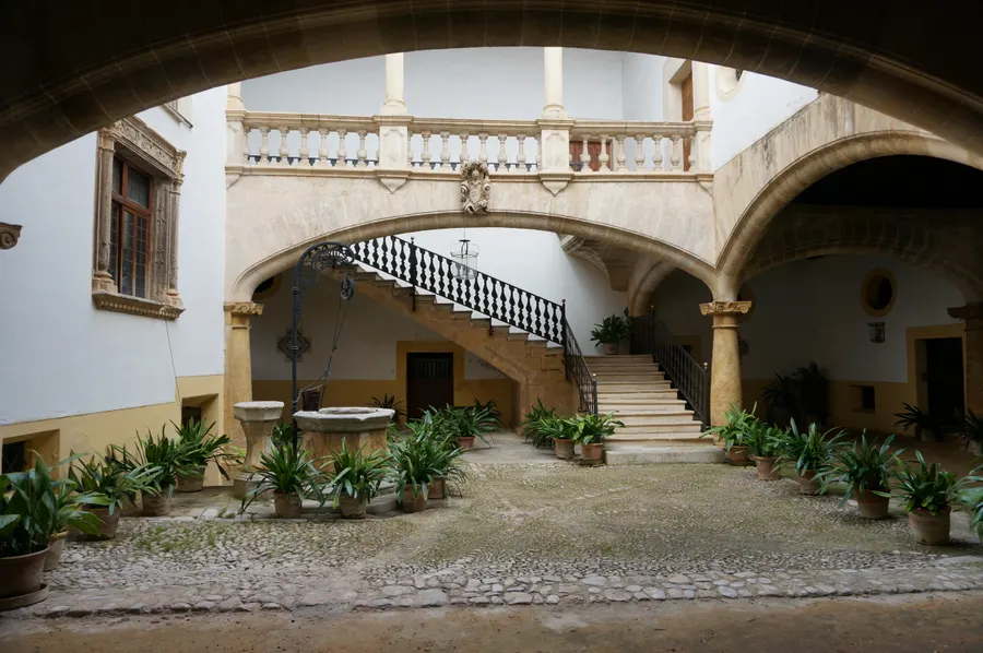 Innenhof eines Stadtpalastes in Palma mit Rundbögen, steinerner Freitreppe, schmiedeeisernem Geländer und Topfpflanzen auf Kopfsteinpflaster.