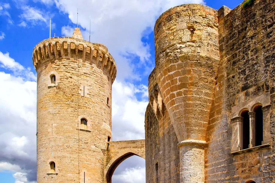 Untersicht auf zwei Türme des Castell de Bellver, dazwischen ein Torbogen, vor blauem Himmel mit Wolken.