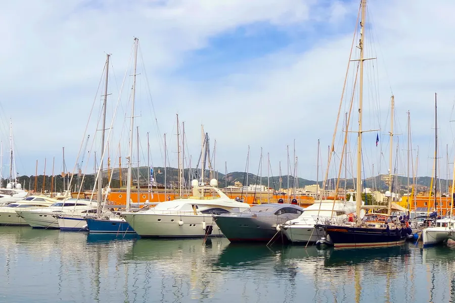 Motor- und Segelboote nebeneinander an einem Steg im Hafen von Palma, dahinter die Stadtsilhouette und Hügel.
