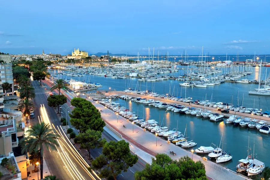 Blick von oben auf den Yachthafen von Palma in der Abenddämmerung, mit Lichtspuren auf der Uferstraße und der Kathedrale am Horizont.
