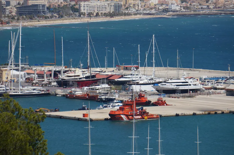 Hafenbecken von Palma mit Segelbooten und einem roten Rettungsboot, dahinter die Küstenlinie der Stadt und Hügel am Horizont.