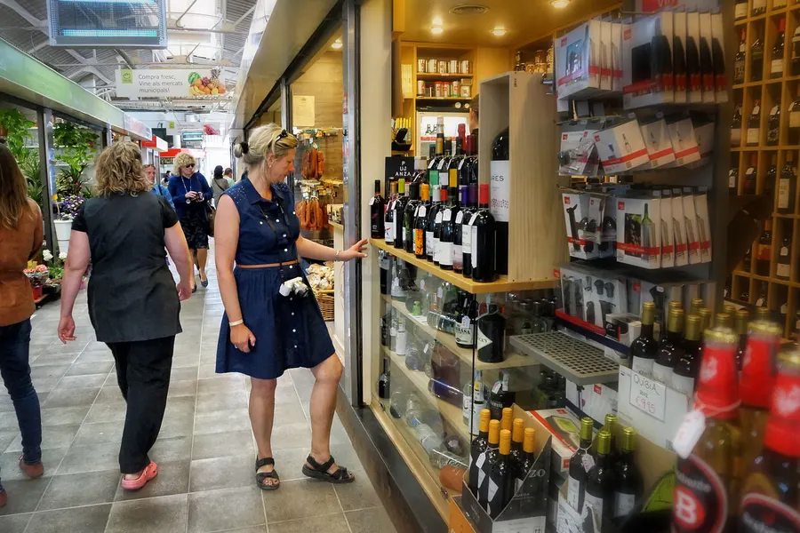 Gang im Mercat de Santa Catalina mit einem Weinstand, Regalen voller Flaschen und zwei Kundinnen davor.
