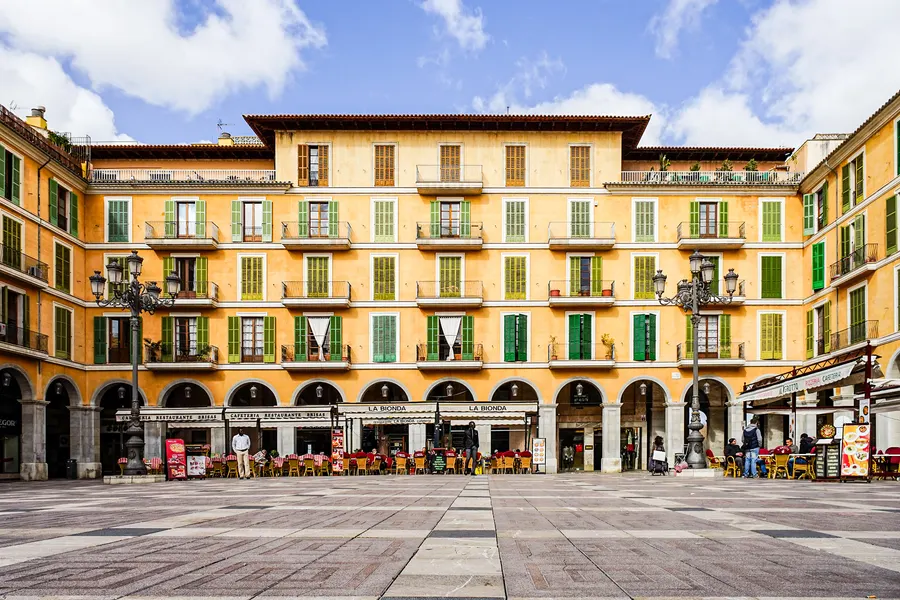 Die Plaça Major in Palma mit ockergelben Arkadenhäusern, grünen Fensterläden und Straßencafés unter den Bögen.