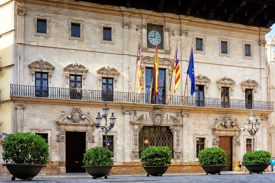 Die Fassade des Rathauses von Palma mit weit vorspringendem Holzdach, Uhr und drei Fahnen.
