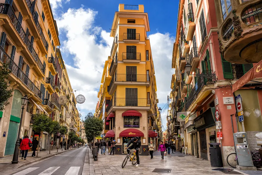Belebte Geschäftsstraße in Palma mit hohen Wohnhäusern, Markisen, Radfahrern und Passanten.
