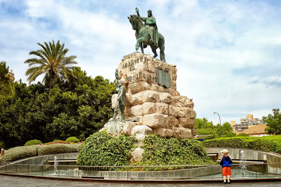 Bronzenes Reiterstandbild auf einem Felssockel in einer Parkanlage in Palma, umgeben von Hecken und Palmen.
