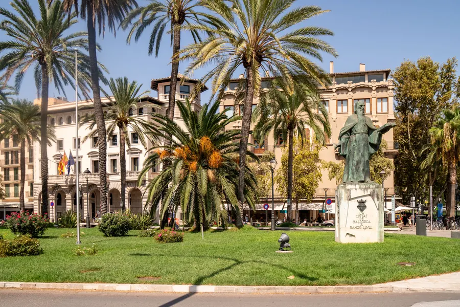 Das Denkmal für Ramon Llull in einer Grünanlage in Palma, umgeben von Palmen und Rasen, dahinter Häuserfronten.
