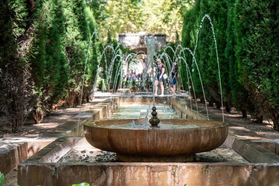 Steinbrunnen mit Wasserfontänen in den Gärten von S'Hort del Rei in Palma, gesäumt von hohen geschnittenen Hecken.
