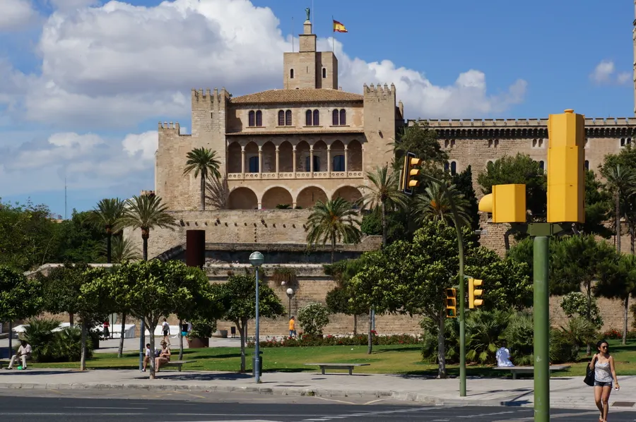 Der Palau de l'Almudaina in Palma, ein wehrhafter Palastbau aus hellem Stein mit Zinnen und Arkaden, davor Palmen und eine breite Straße.
