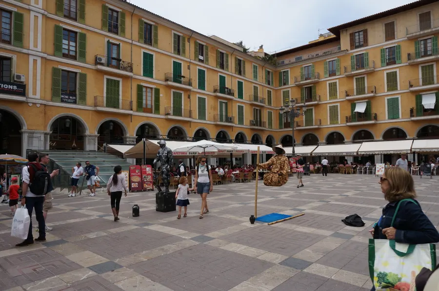 Die Plaça Major in Palma, ein rechteckiger Platz mit ockergelben Arkadenhäusern, Sonnenschirmen von Straßencafés und Passanten.