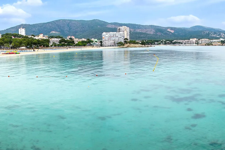 Weite Fläche hellen, flachen Wassers vor Palmanova mit Bojenkette, dahinter der Strand, Hotelbauten und die Berge.