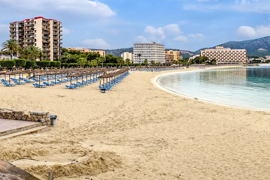 Reihen zusammengeklappter Liegen und Schirme am Strand von Palmanova am frühen Morgen, dahinter Hotels und Palmen.